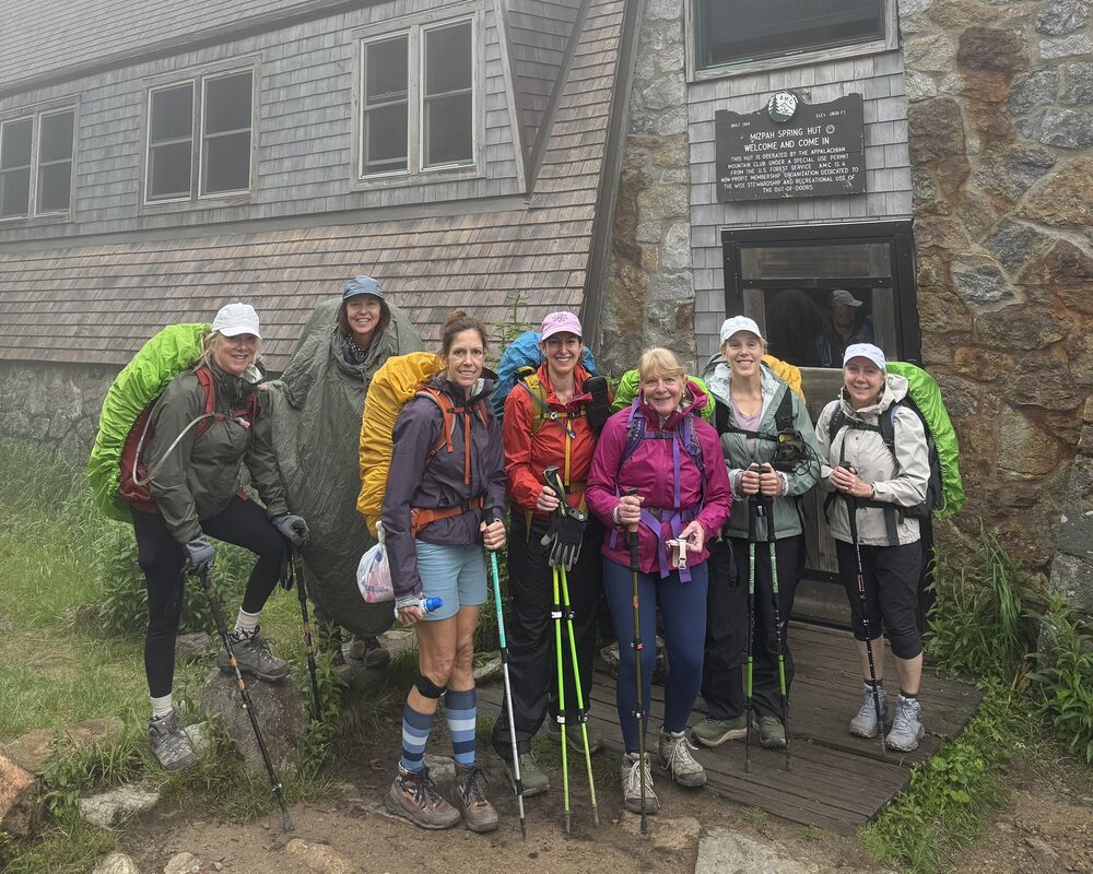 A group of hikers with backpacks and trekking poles gathered outside a stone mountain hut