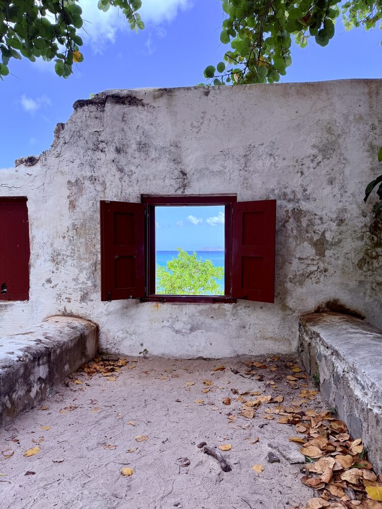 A red-shuttered window in an old whitewashed stone ruin, framing green leaves and blue sea beyond
