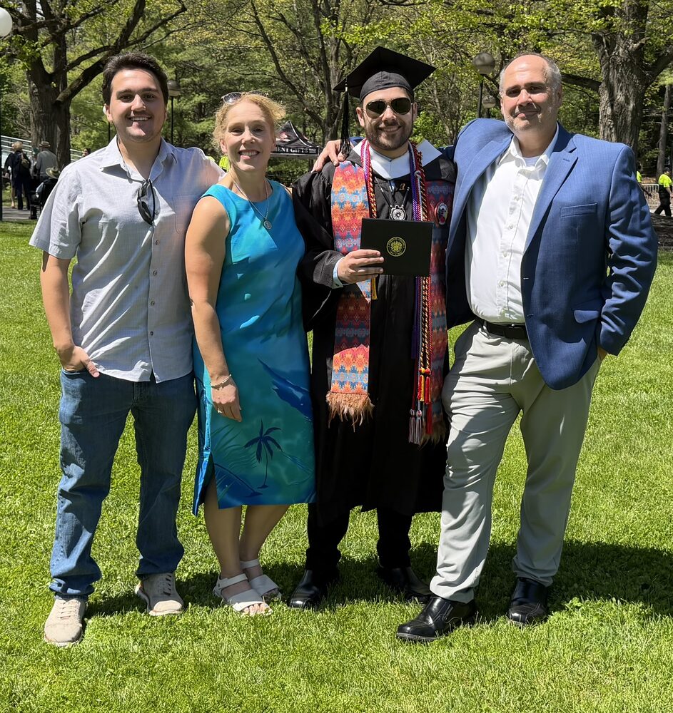 Family of four on a lawn at Will's college graduation, the graduate in cap, gown, and rainbow stole holding his diploma